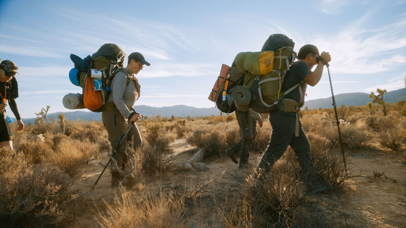 The image shows a group of hikers trekking through a dry, grassy landscape under a bright sky. They are heavily laden with large backpacks, suggesting a multi-day trip. The hikers use trekking poles to aid their journey across the uneven terrain. The environment appears arid, with sparse vegetation and distant hills visible in the background.